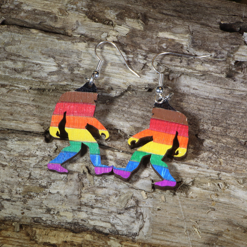 Wooden Bigfoot silhouette earrings painted in rainbow stripes, resting on a wood-grain surface