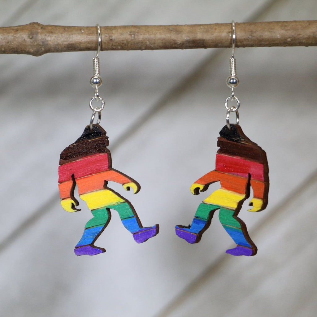 Rainbow-striped wooden Bigfoot earrings hanging from silver ear wires on a wooden branch