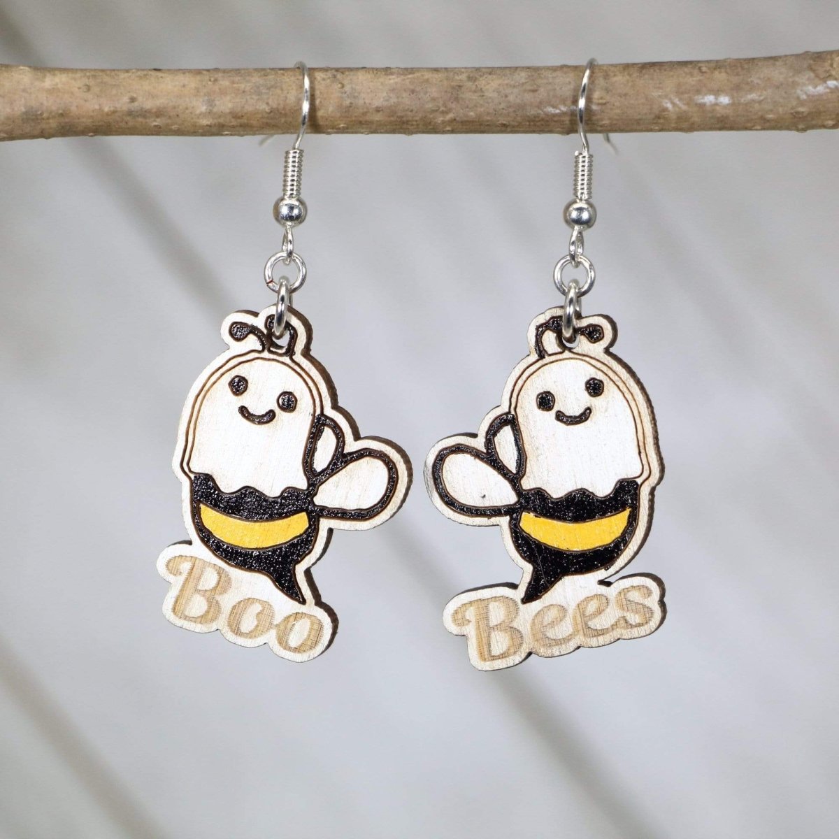 Boo Bees ghost-bee wooden earrings hanging from silver ear wires on a wooden branch