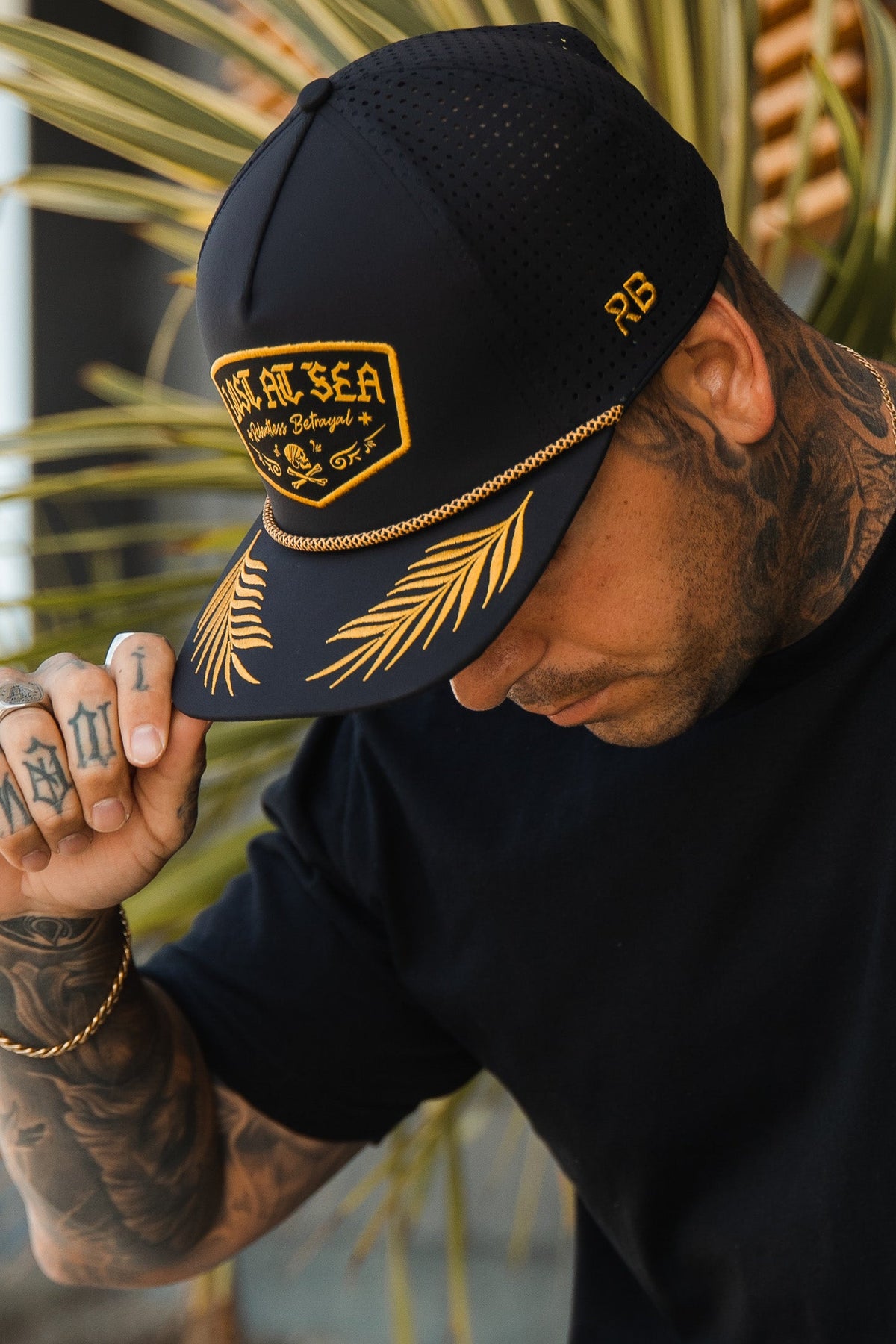 Lost At Sea BLACKOUT Snapback: black cap with gold-colored LOST AT SEA patch, rope trim and leaf embroidery on brim, brim being adjusted.