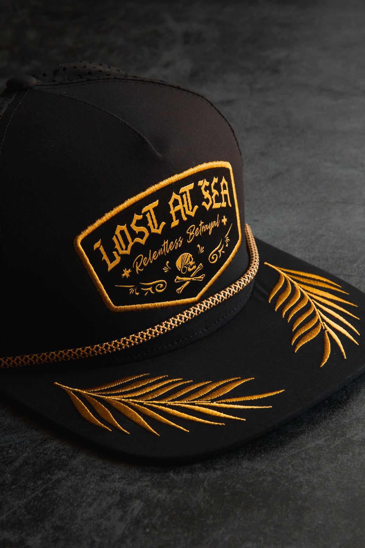 Lost At Sea BLACKOUT Snapback: black cap with gold-colored LOST AT SEA patch, rope trim and leaf embroidery on brim, patch and brim close-up.