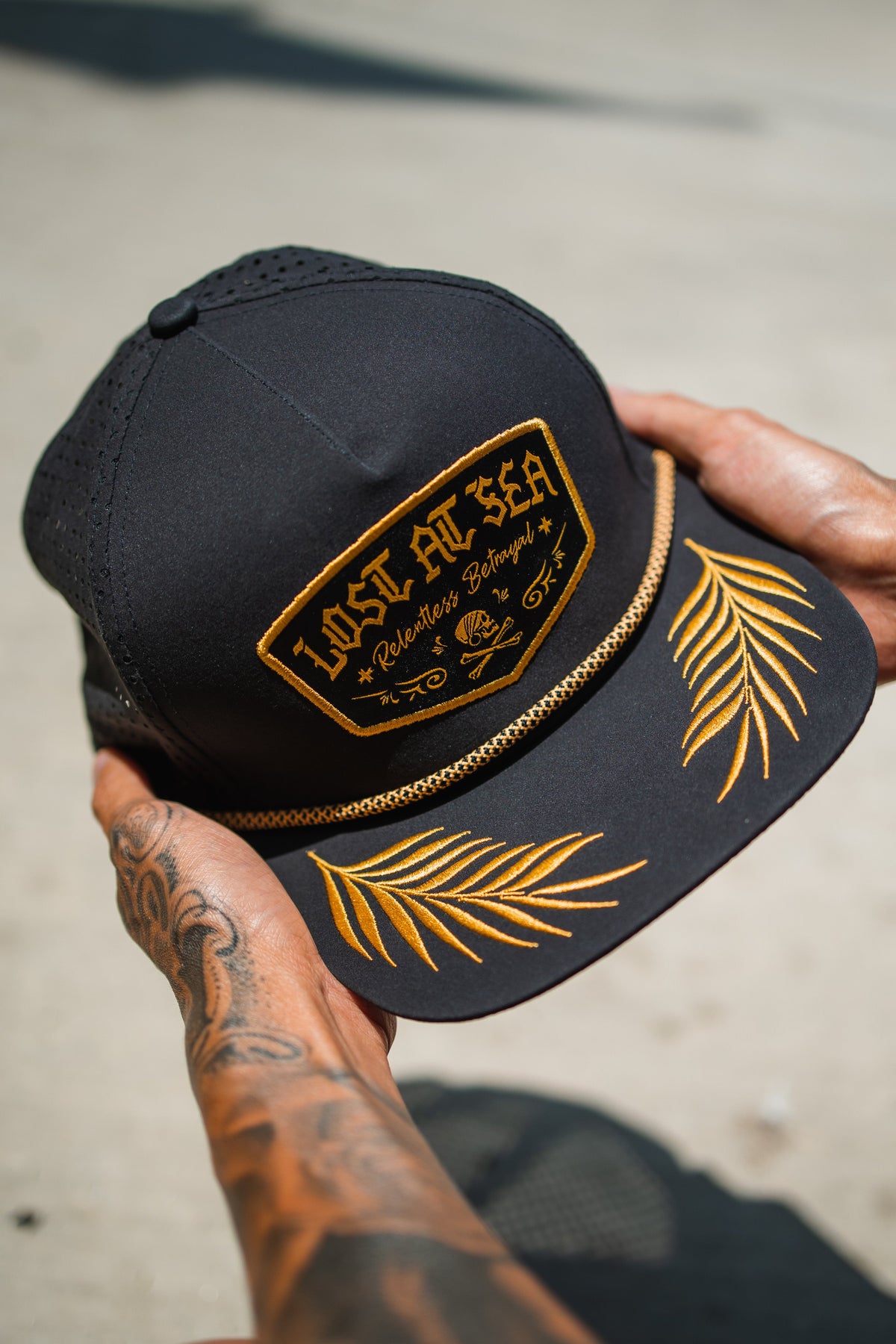Lost At Sea BLACKOUT Snapback: black cap with gold-colored LOST AT SEA patch, rope trim and leaf embroidery on brim, held in hands.