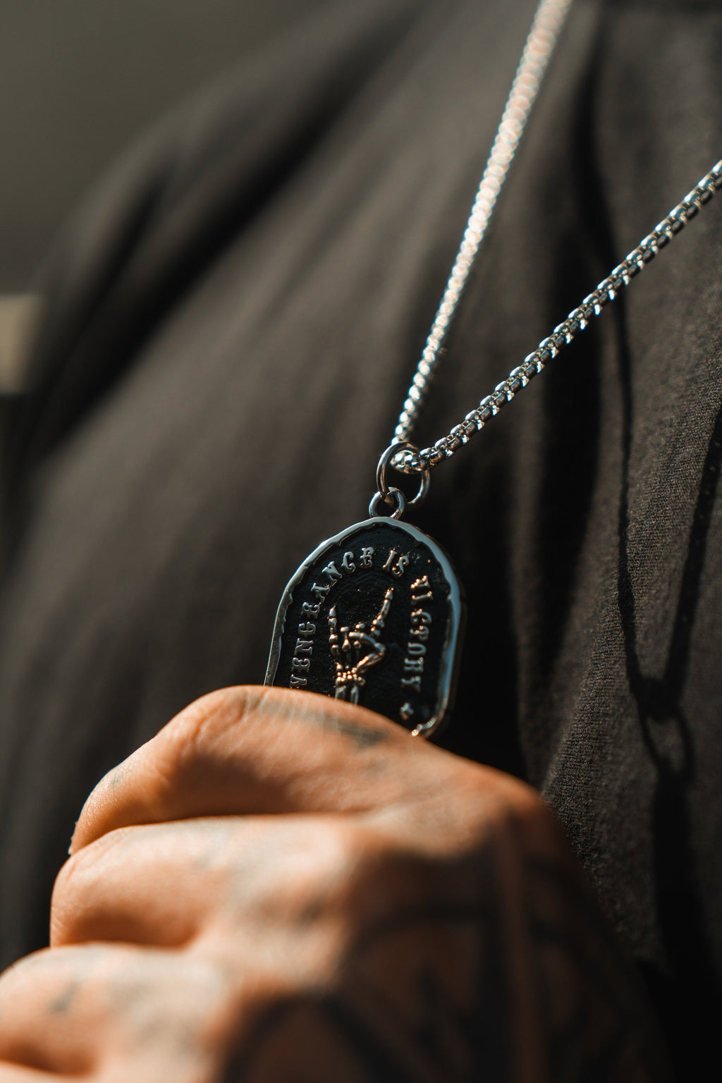 Vengeance Necklace: black shield-shaped pendant with skeletal hand and VENGEANCE IS VICTORY lettering, worn against a dark shirt.