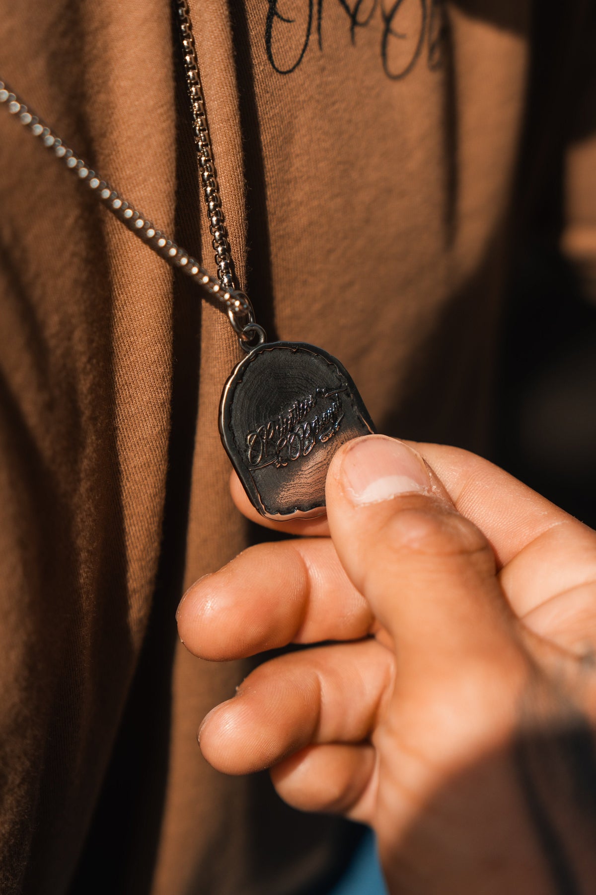 Vengeance Necklace: brand-script reverse on a shield-shaped pendant, reverse held against a shirt.