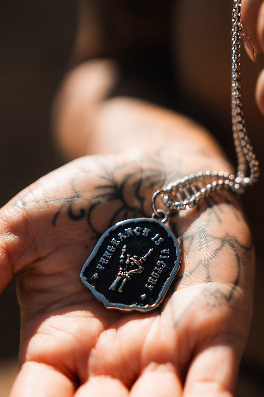 Vengeance Necklace: black shield-shaped pendant with skeletal hand and VENGEANCE IS VICTORY lettering, front held in a hand.