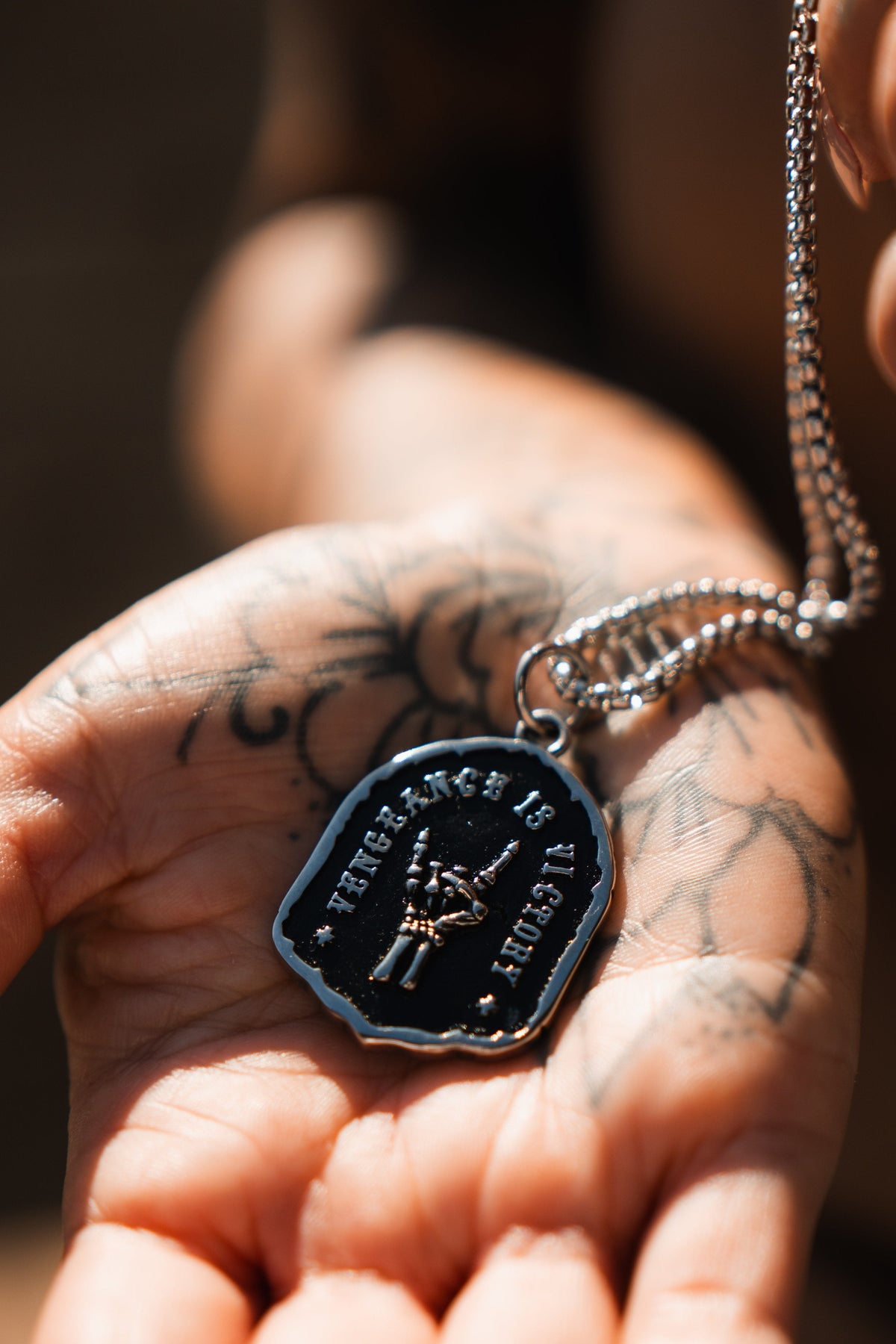 Vengeance Necklace: black shield-shaped pendant with skeletal hand and VENGEANCE IS VICTORY lettering, front held in a hand.