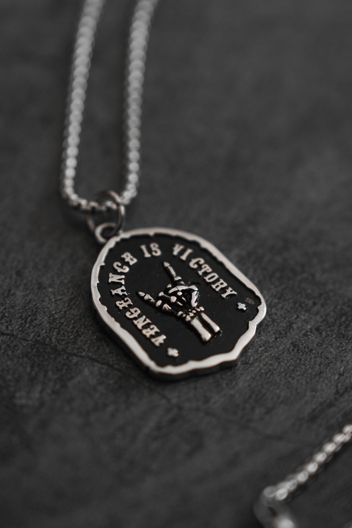 Vengeance Necklace: black shield-shaped pendant with skeletal hand and VENGEANCE IS VICTORY lettering, angled front product view.