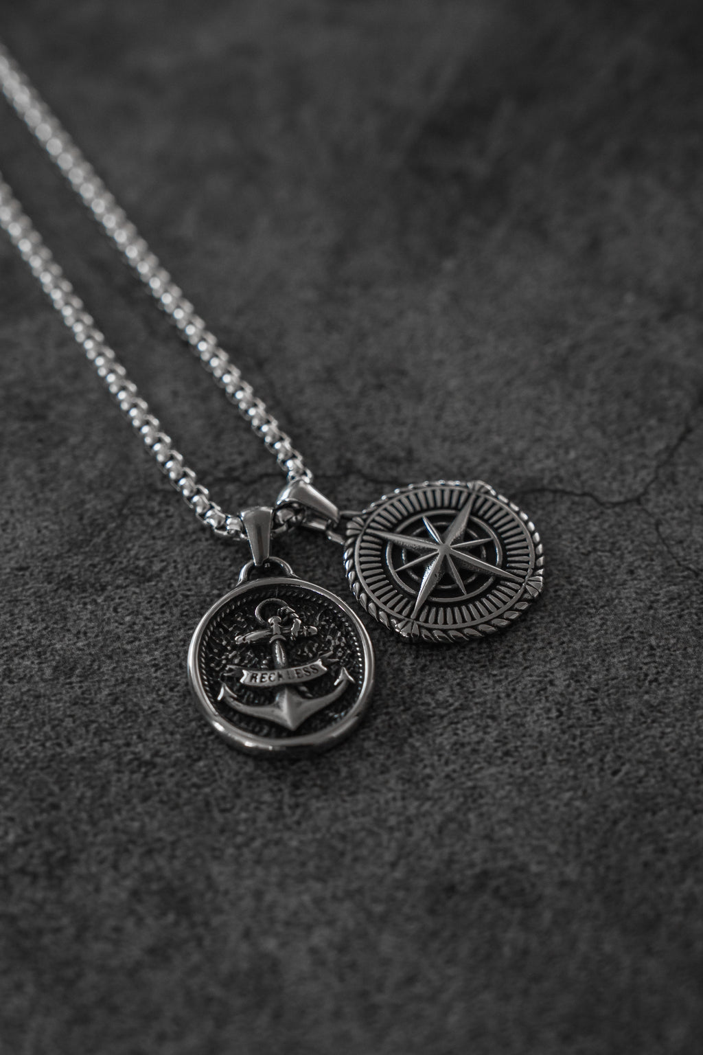 Reckless Soul Necklace: paired round anchor and compass-rose pendants on a silver-tone chain, both pendant faces on gray surface.