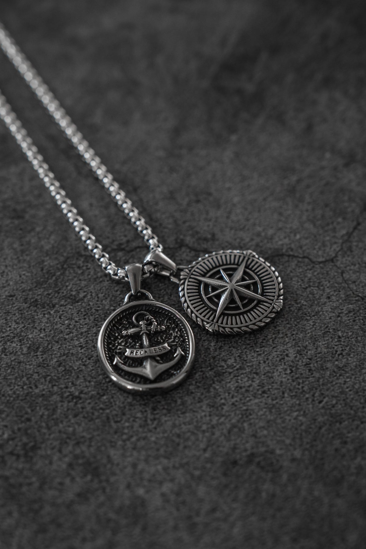 Reckless Soul Necklace: paired round anchor and compass-rose pendants on a silver-tone chain, both pendant faces on gray surface.