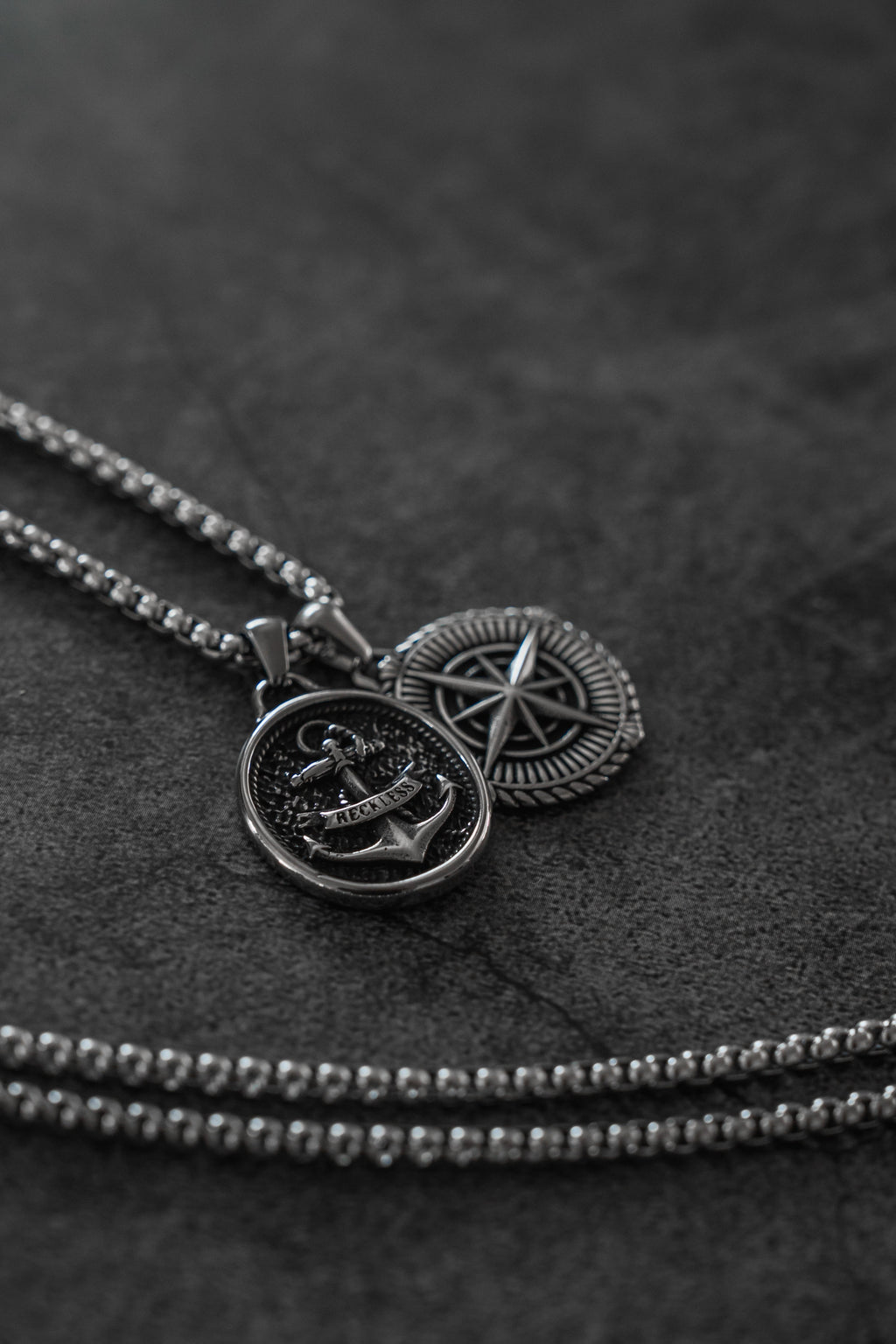 Reckless Soul Necklace: paired round anchor and compass-rose pendants on a silver-tone chain, pendants and chain on gray surface.