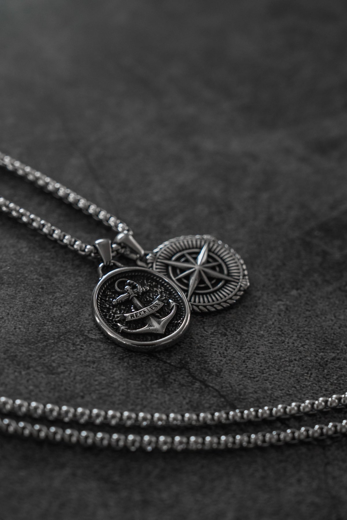Reckless Soul Necklace: paired round anchor and compass-rose pendants on a silver-tone chain, pendants and chain on gray surface.