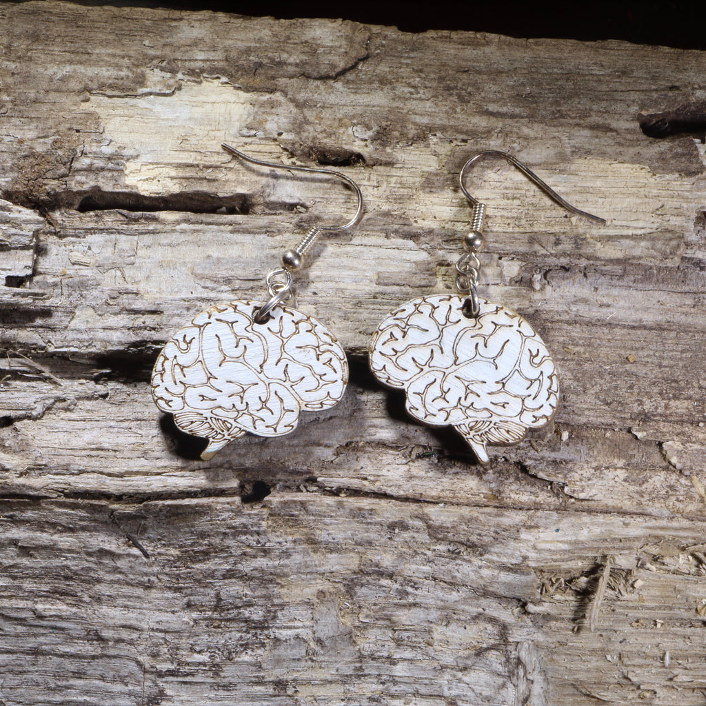 Wood-etched brain silhouette earrings hanging from silver fishhook wires, on a weathered wood-grain surface