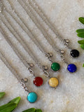 Six Stone Charm Necklace colorways laid out together — sky blue, green, red, yellow, black, and blue stone pendants on chains