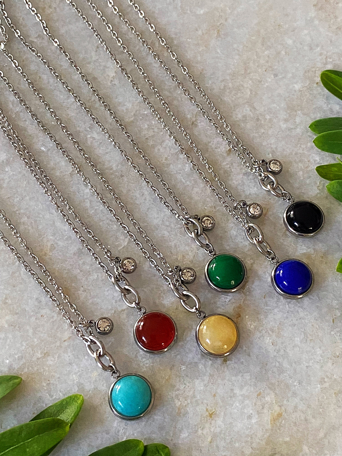 Six Stone Charm Necklace colorways laid out together — sky blue, green, red, yellow, black, and blue stone pendants on chains