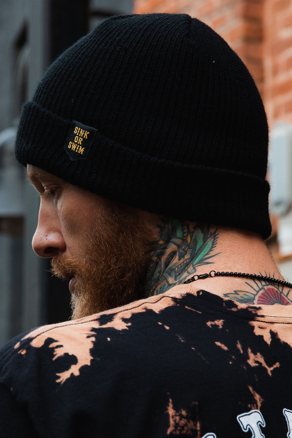 Sink or Swim Beanie: black rib-knit cuffed beanie with small gold-colored SINK OR SWIM lettering, rear-side view on a model.