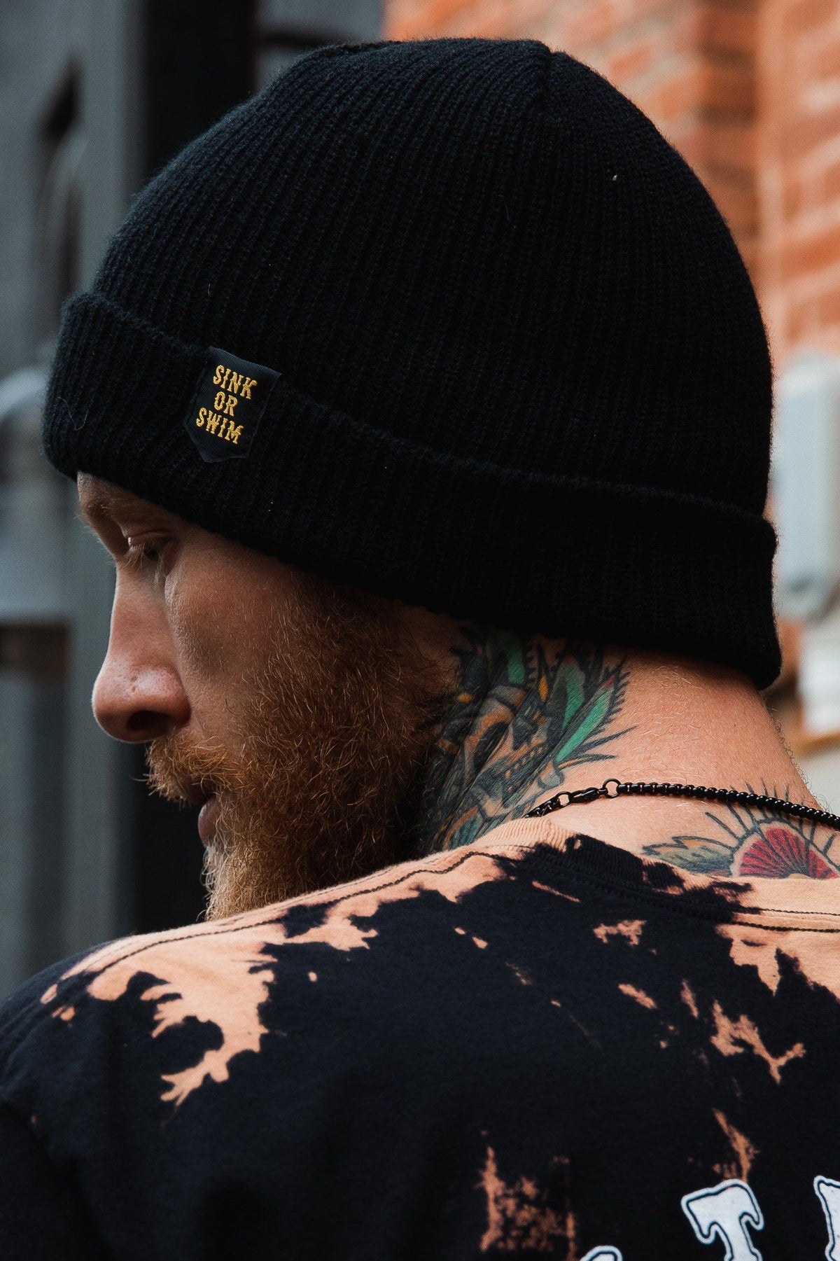 Sink or Swim Beanie: black rib-knit cuffed beanie with small gold-colored SINK OR SWIM lettering, rear-side view on a model.