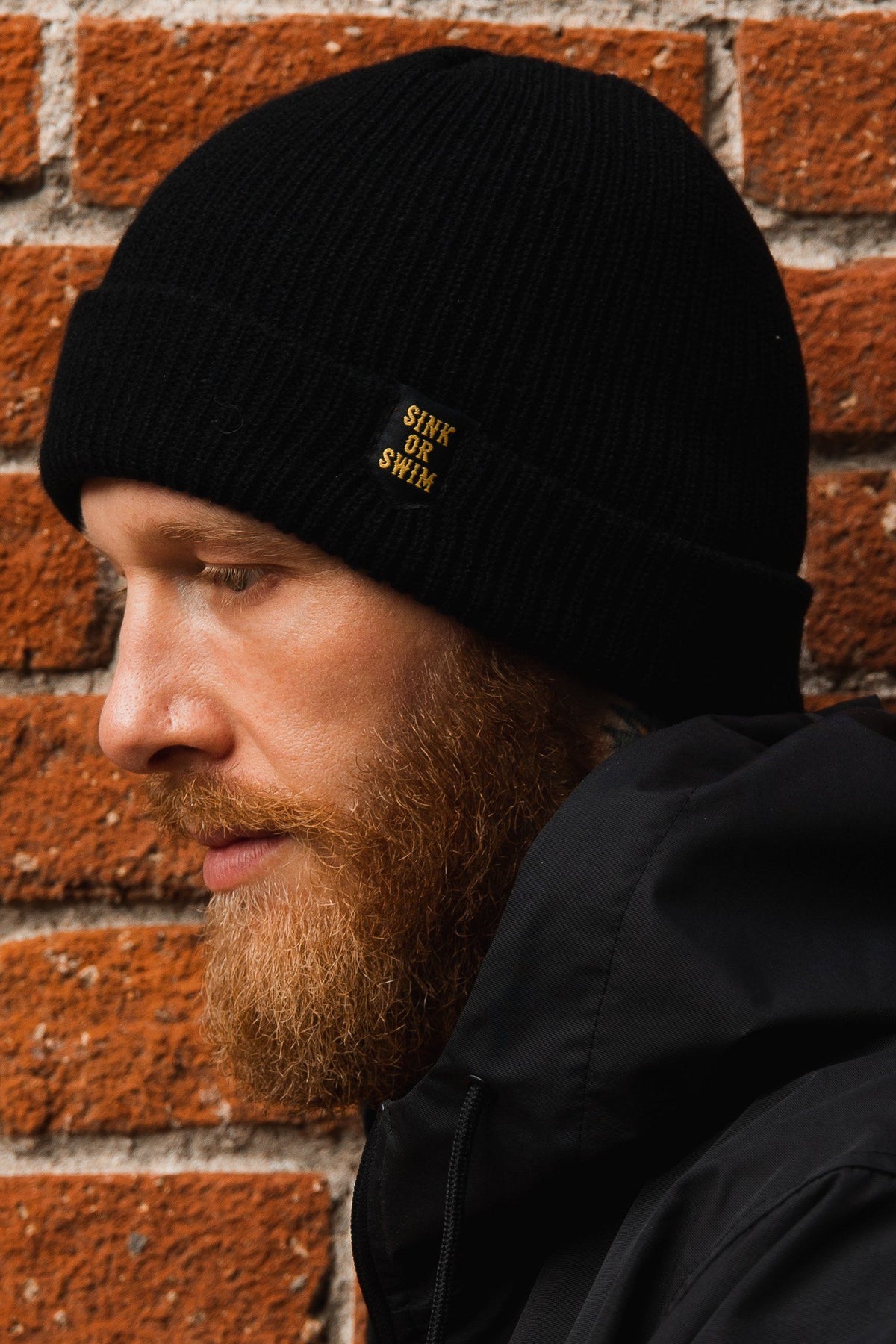 Sink or Swim Beanie: black rib-knit cuffed beanie with small gold-colored SINK OR SWIM lettering, side view on a model.