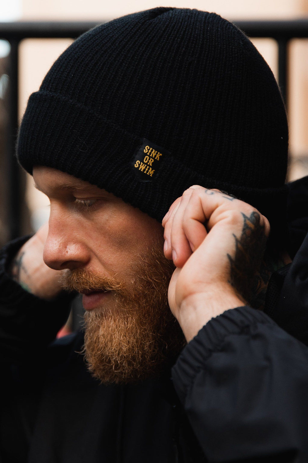 Sink or Swim Beanie: black rib-knit cuffed beanie with small gold-colored SINK OR SWIM lettering, being adjusted on a model.