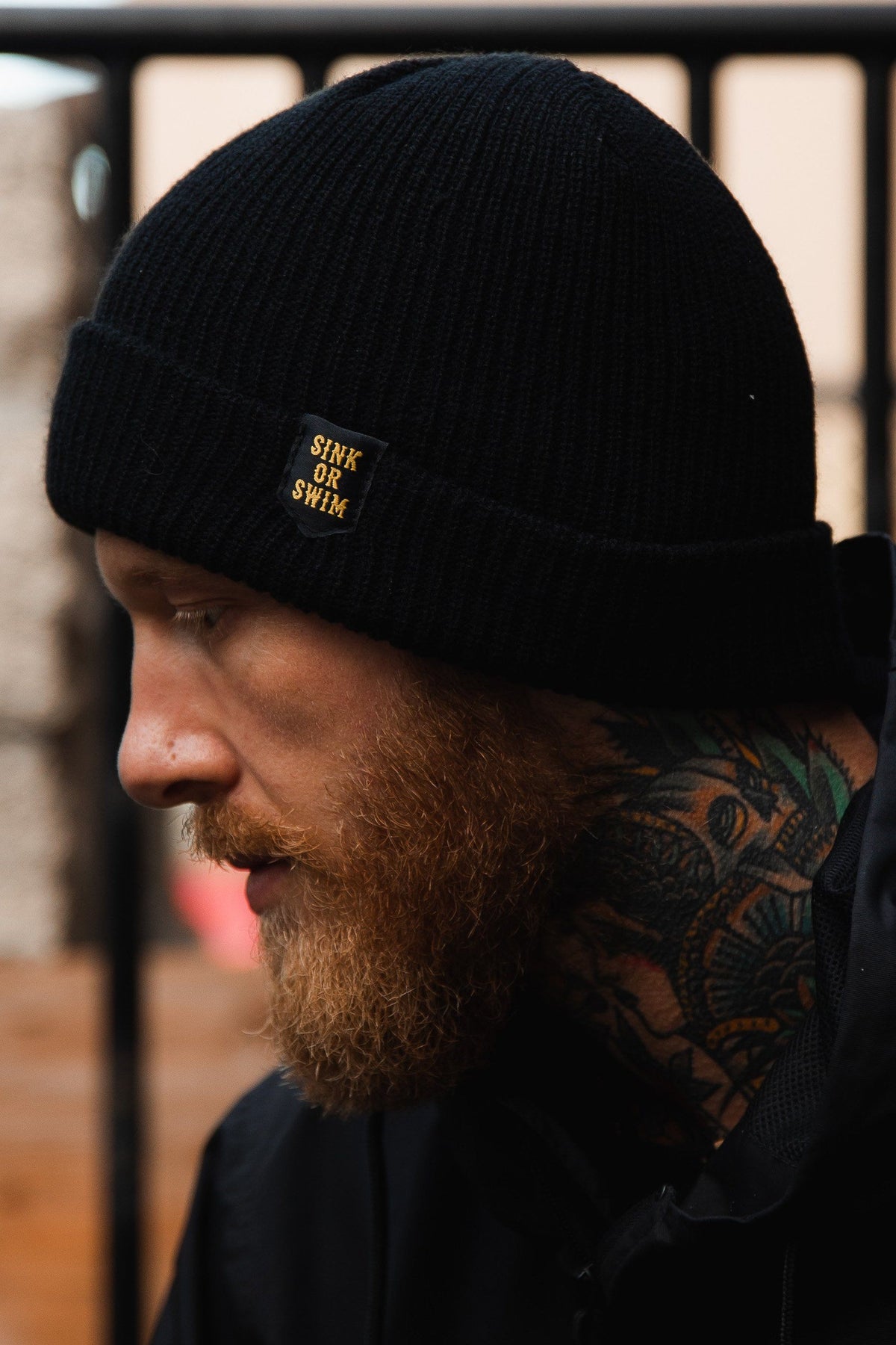 Sink or Swim Beanie: black rib-knit cuffed beanie with small gold-colored SINK OR SWIM lettering, side view on a model.