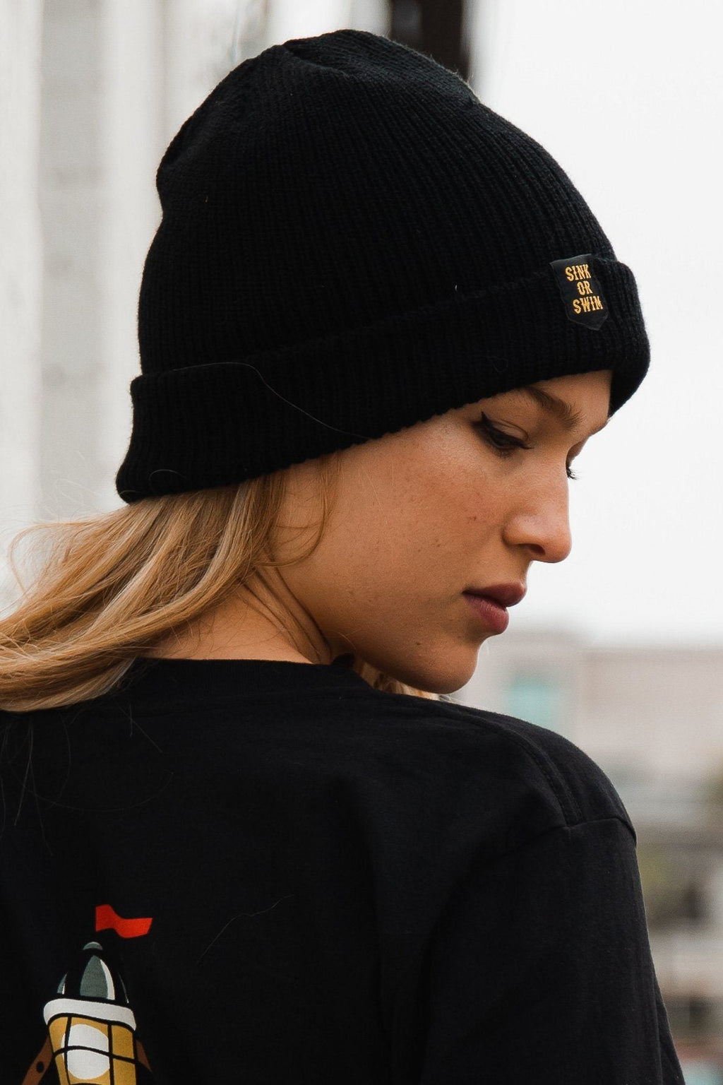 Sink or Swim Beanie: black rib-knit cuffed beanie with small gold-colored SINK OR SWIM lettering, side view on a model.