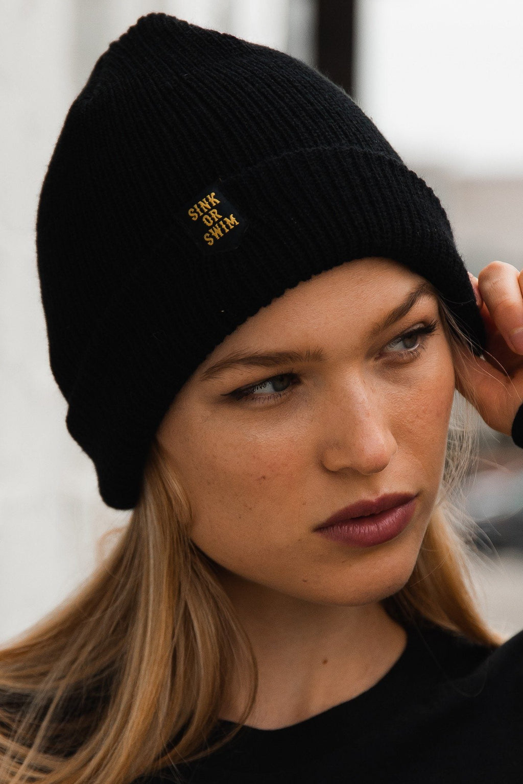Sink or Swim Beanie: black rib-knit cuffed beanie with small gold-colored SINK OR SWIM lettering, angled view on a model.