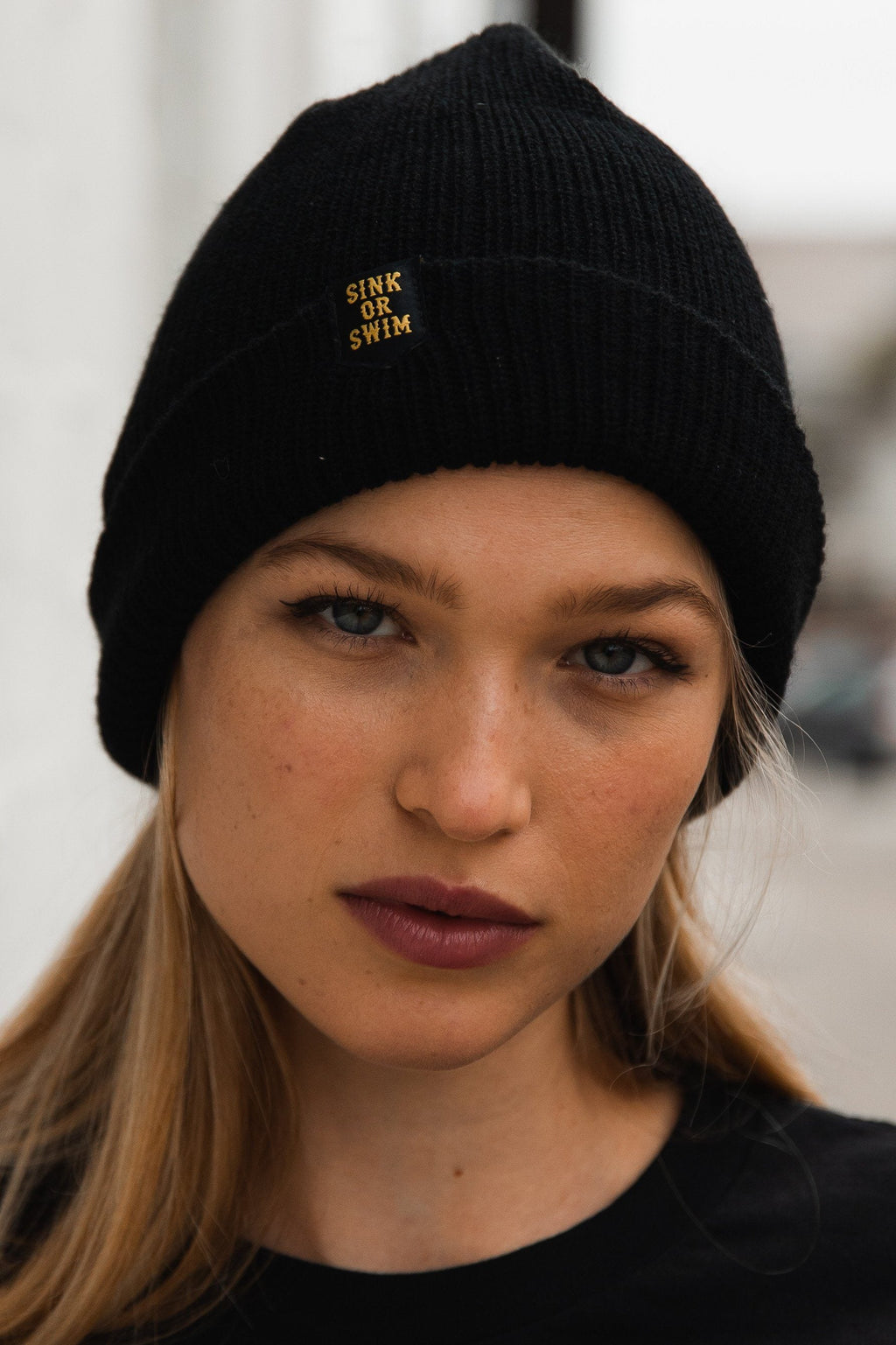 Sink or Swim Beanie: black rib-knit cuffed beanie with small gold-colored SINK OR SWIM lettering, front view on a model.