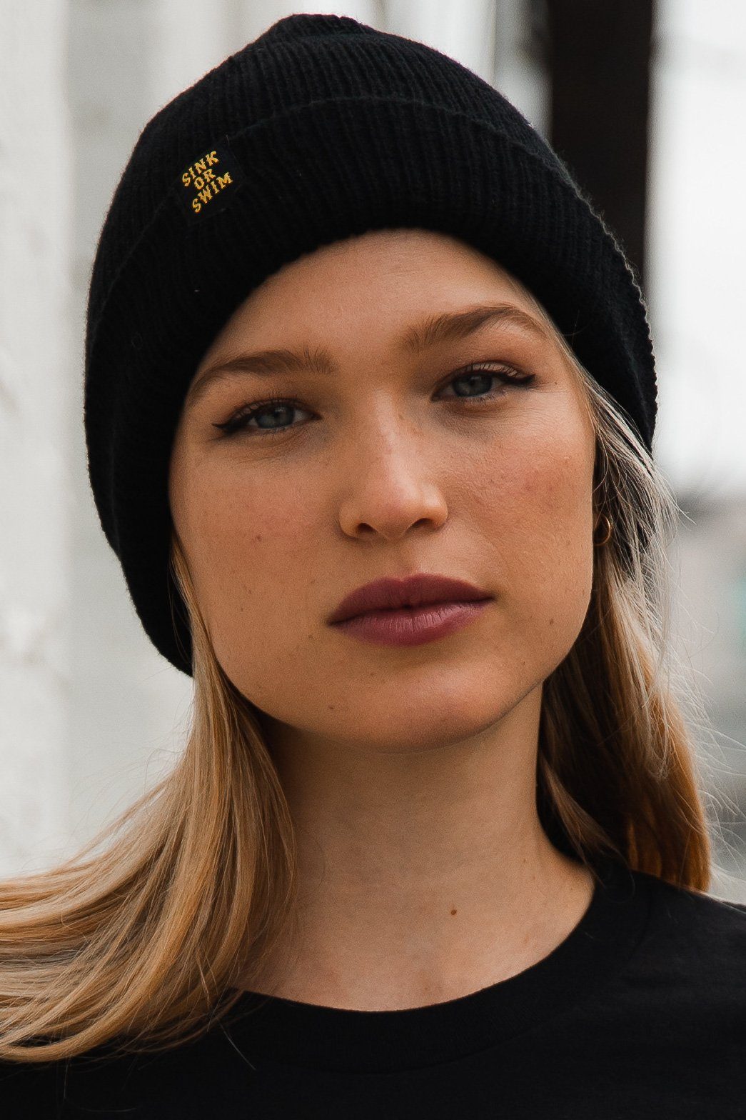 Sink or Swim Beanie: black rib-knit cuffed beanie with small gold-colored SINK OR SWIM lettering, front view on a model.