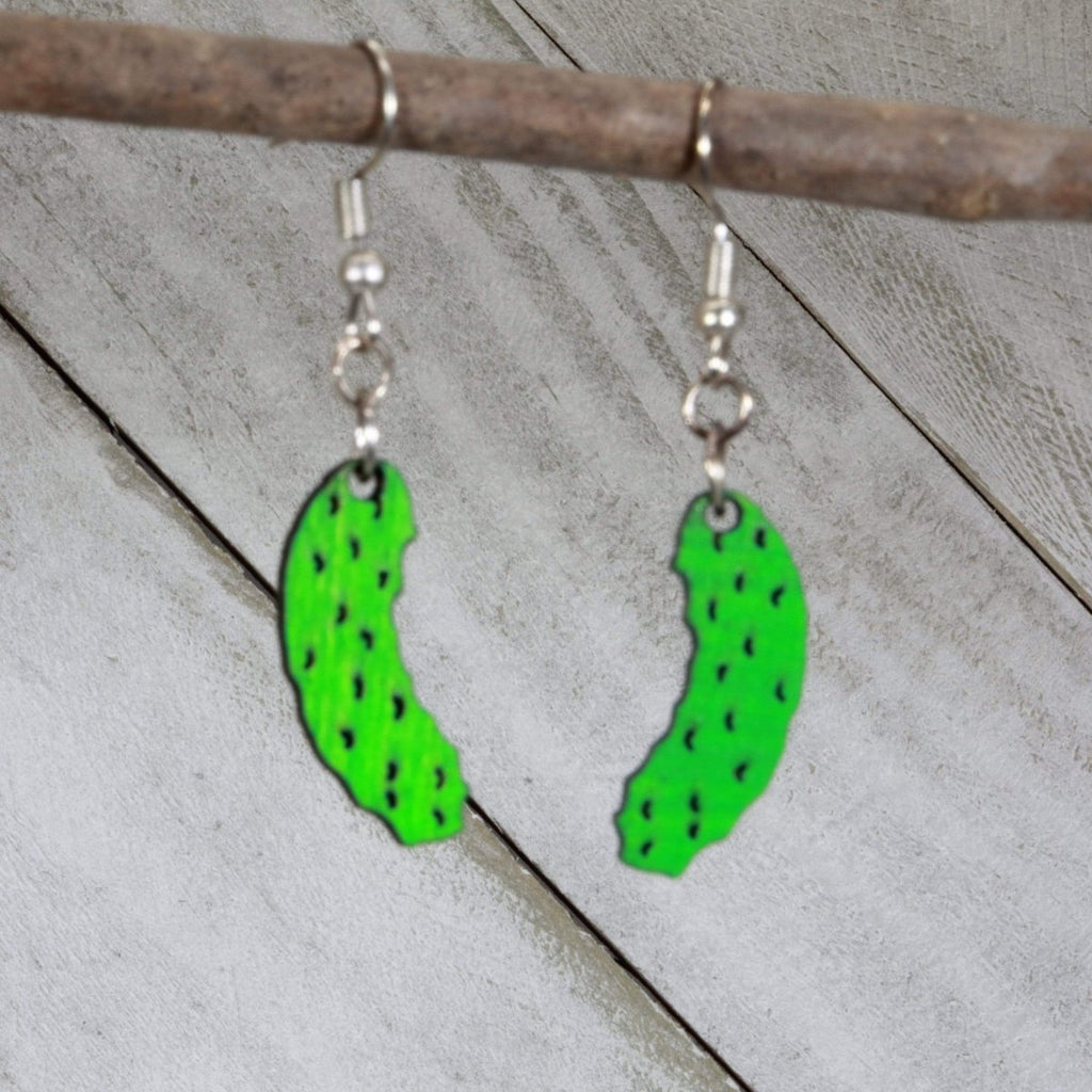 Green pickle-shaped wooden earrings hanging from silver ear wires on a wooden branch