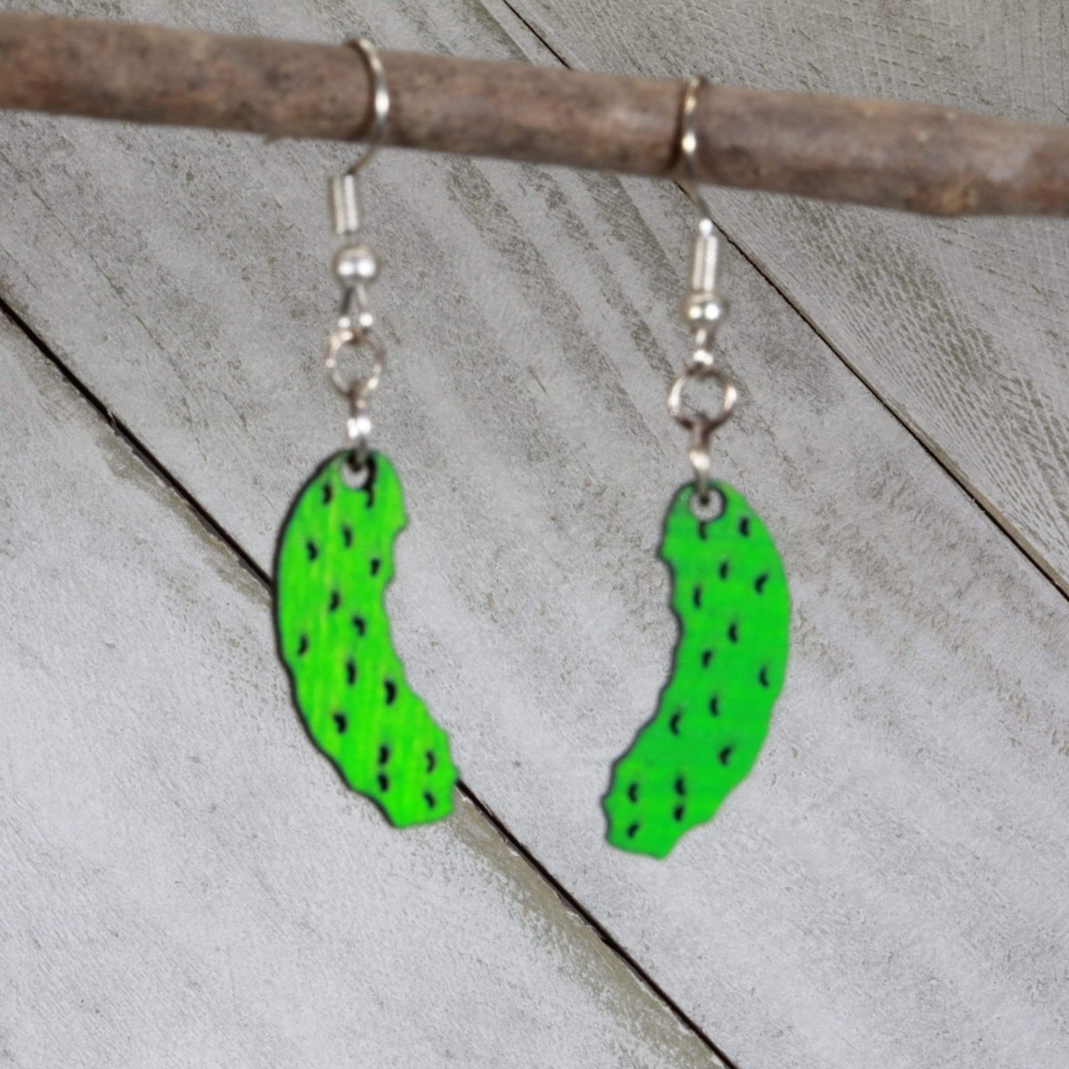 Green pickle-shaped wooden earrings hanging from silver ear wires on a wooden branch
