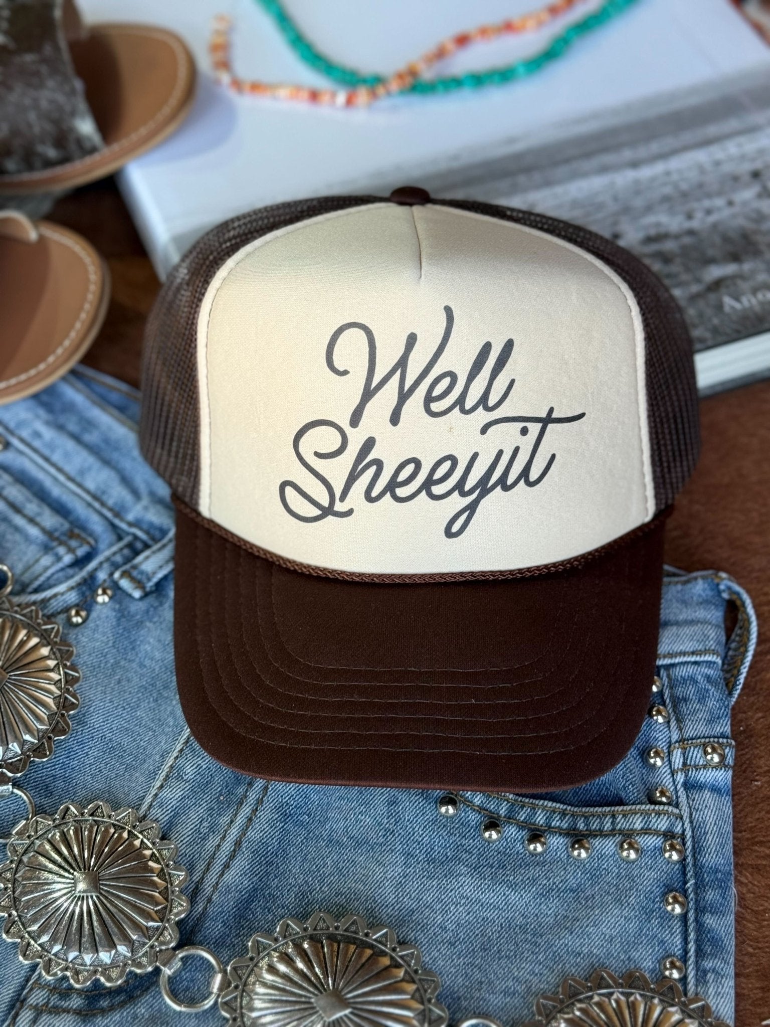 Brown and tan Well Sheeyit trucker hat styled flat with denim and jewelry accessories