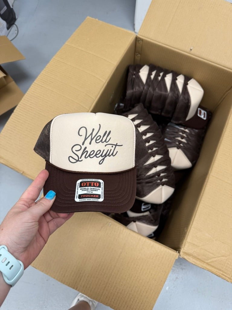 Brown and tan trucker hat with cursive Well Sheeyit script, mesh back, held above an open shipping box